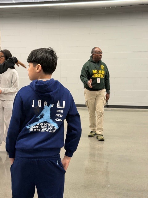 Three individuals stand in a large indoor space with concrete floors and white brick walls. One person wears a green sweatshirt and appears to be instructing; another wears a blue tracksuit with motivational text on the back.