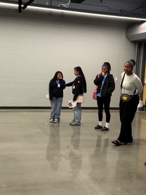 Four individuals stand against a gray cinder‑block wall, holding papers or booklets. They are dressed casually and facing the camera.