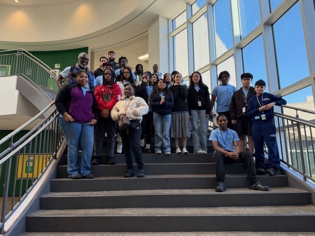 A group of Mills Middle School 8th‑grade AVID students and a staff member pose together on a wide staircase inside a bright, modern school building with large windows.