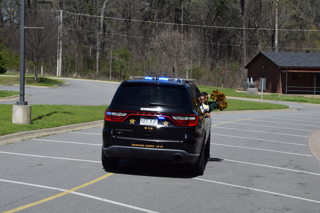 Two students lean out of a sheriff's office patrol car. They are cheering with pom poms.