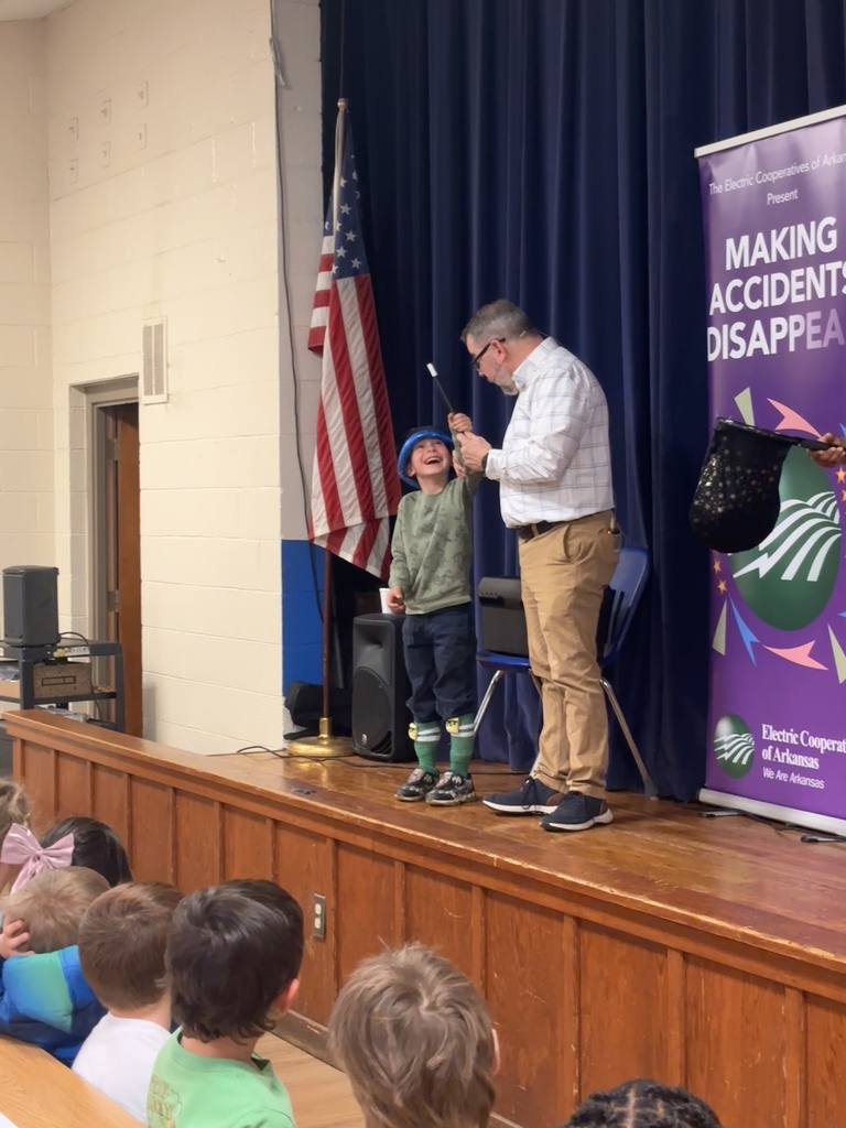 Scott Davis in front our Lawson Students explaining what his show is all about (electrical safety). He is on stage with a banner behind him that reads "making accidents disappear". He has a student helper who is wearing a green "lucky me" shirt.  He has a second student helper who is in kindergarten. He is wearing a green camo shirt and now has a magicians hat and wand. 