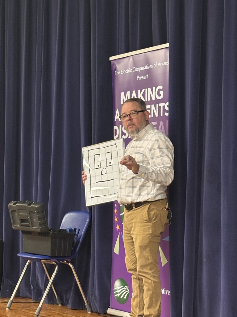Scott Davis in front our Lawson Students explaining what his show is all about (electrical safety). He is on stage with a banner behind him that reads "making accidents disappear". He is holding a small dry erase board with an electrical outlet drawn on it named "Sparky". 