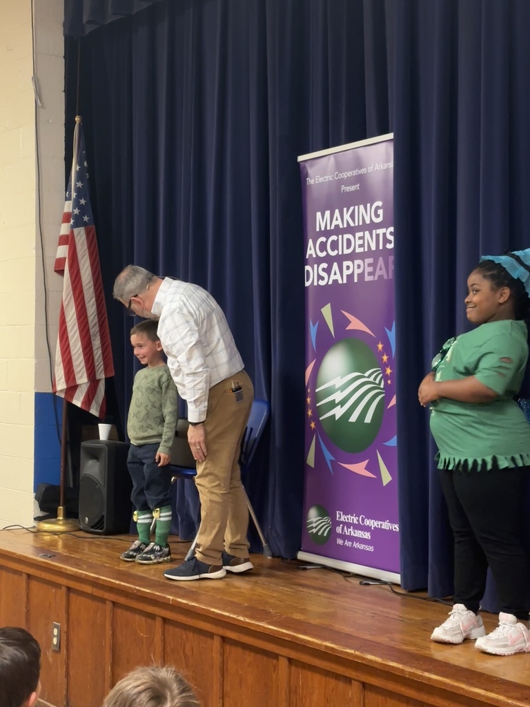 Scott Davis in front our Lawson Students explaining what his show is all about (electrical safety). He is on stage with a banner behind him that reads "making accidents disappear". He has a student helper who is wearing a green "lucky me" shirt.  He has a second student helper who is in kindergarten. He is wearing a green camo shirt. 