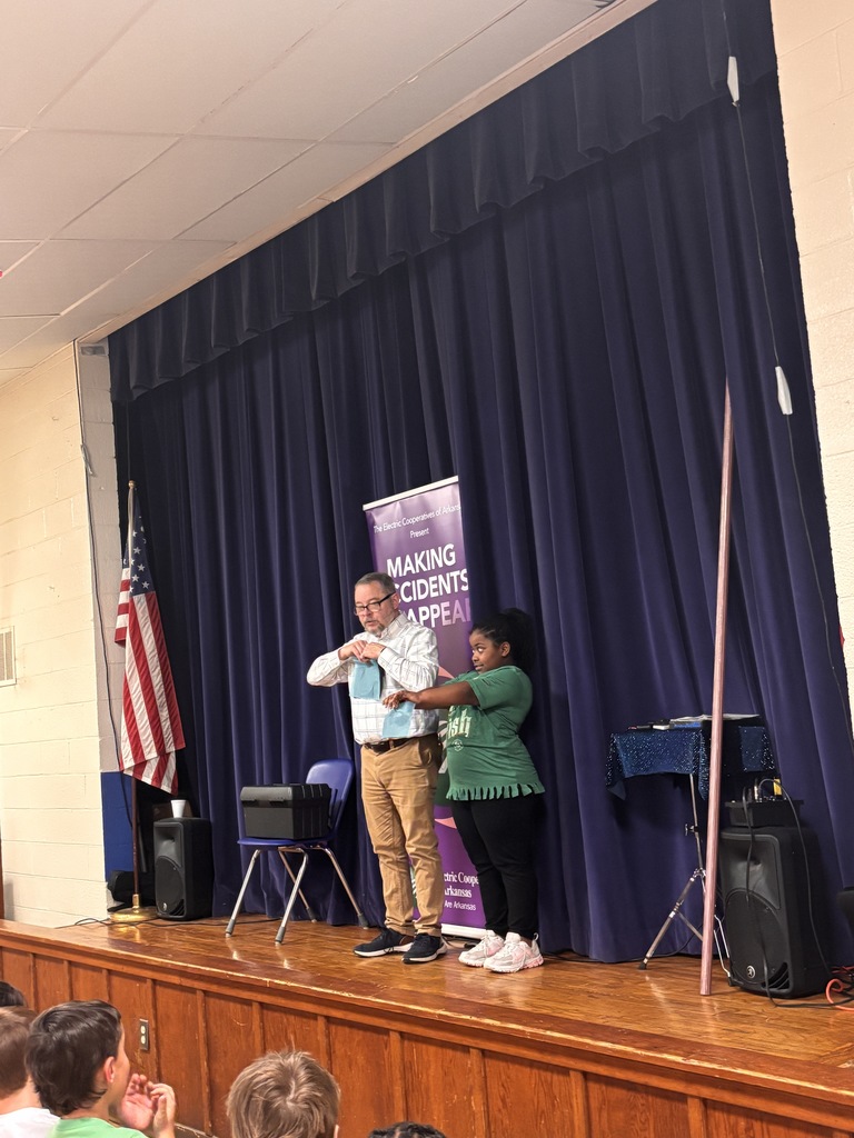 Scott Davis in front our Lawson Students explaining what his show is all about (electrical safety). He is on stage with a banner behind him that reads "making accidents disappear". He has a student helper who is wearing a green "lucky me" shirt.  She is holding out two pieces of tissue paper and being instructed to rip them. 