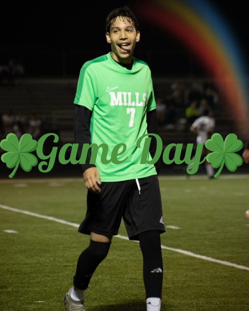 A Mills soccer player wearing a green jersey with the number 7 smiles while walking on the field during a nighttime game. “Game Day” with green shamrocks is overlaid on the image.