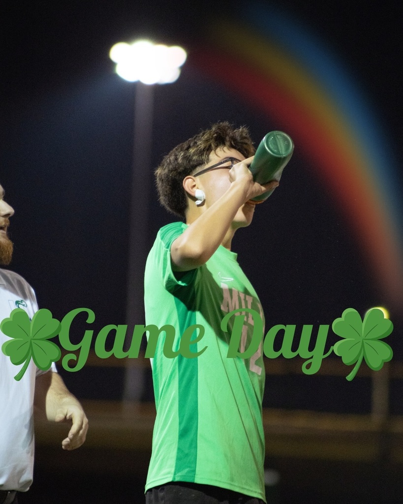 A Mills soccer player wearing jersey drinks from a green water bottle under stadium lights. “Game Day” with shamrocks is overlaid on the image.