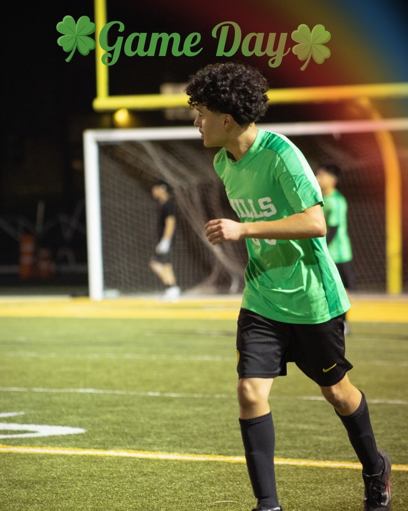 A Mills soccer player wearing jersey number 99 runs on the field during a nighttime game, with “Game Day” and shamrocks overlaid at the top.