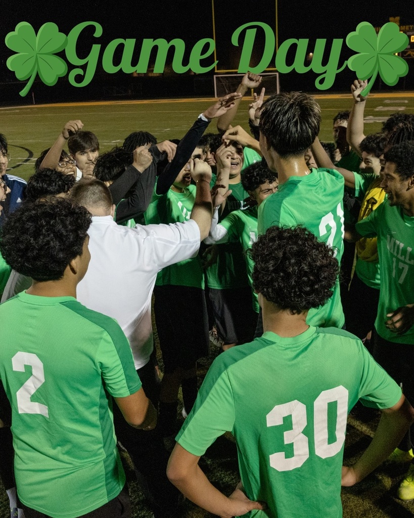 Mills soccer players in green jerseys gather in a pre‑game huddle with a coach, raising their hands together. “Game Day” with shamrocks appears above the group.