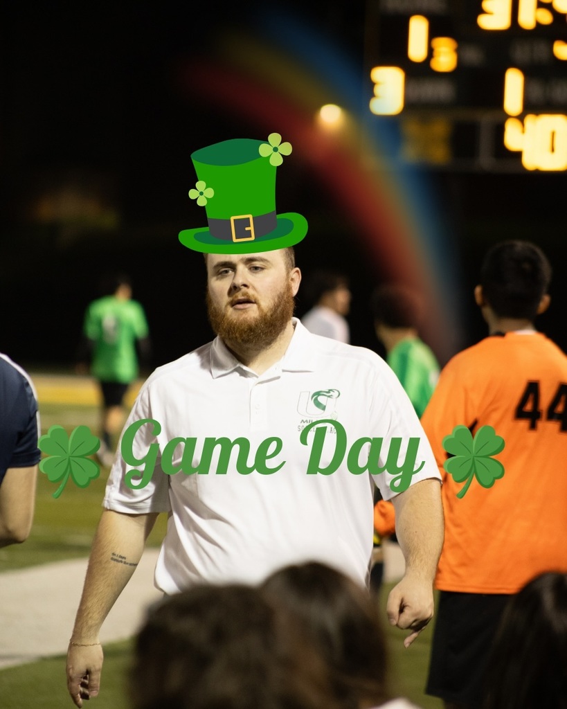 A Mills soccer coach in a white polo stands on the sideline during a game, digitally edited with a green leprechaun hat and “Game Day” text with shamrocks.