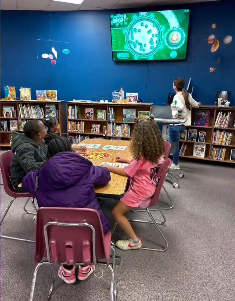 Children sit around a table in the school library playing a winter‑themed bingo game. A woman stands beside a laptop connected to a large screen displaying Bingo!” with numbered balls. Bookshelves, space‑themed decorations, and hanging planets fill the background.