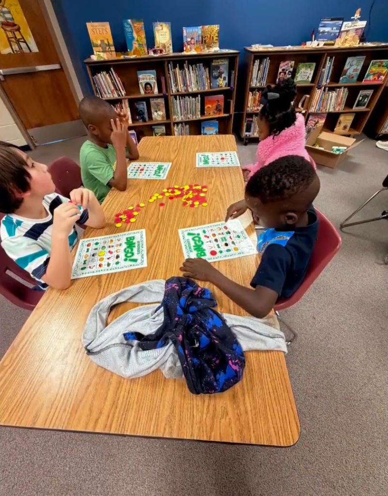 Young children sit around a table in a classroom or library, marking bingo cards with red and yellow chips. Bookshelves filled with children’s books line the background, creating a cozy learning environment.