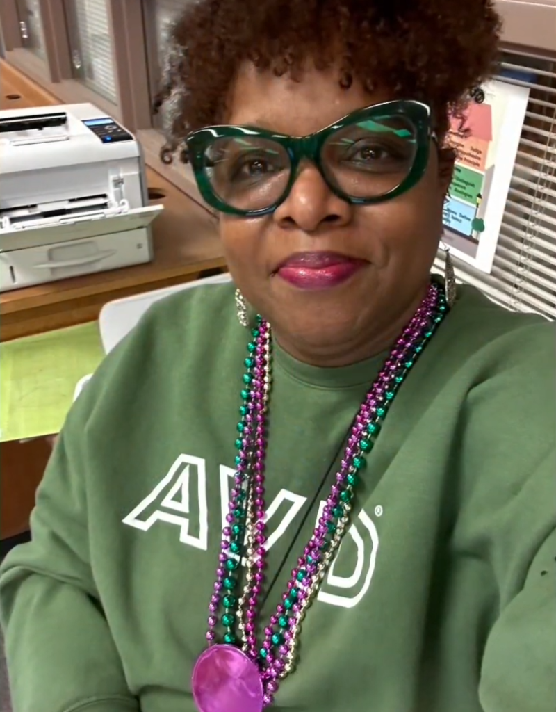 A person wearing a green AVID sweatshirt and colorful Mardi Gras beads smiles in a classroom or office setting. They wear green glasses, and a printer, bulletin board, and desk materials are visible behind them.