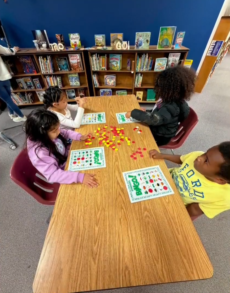 Four children sit at a wooden table in the library playing bingo. Each child has a bingo card and colorful red and yellow chips. Bookshelves filled with children’s books and decorative letters spelling “READ” and “GO!” are visible behind them.