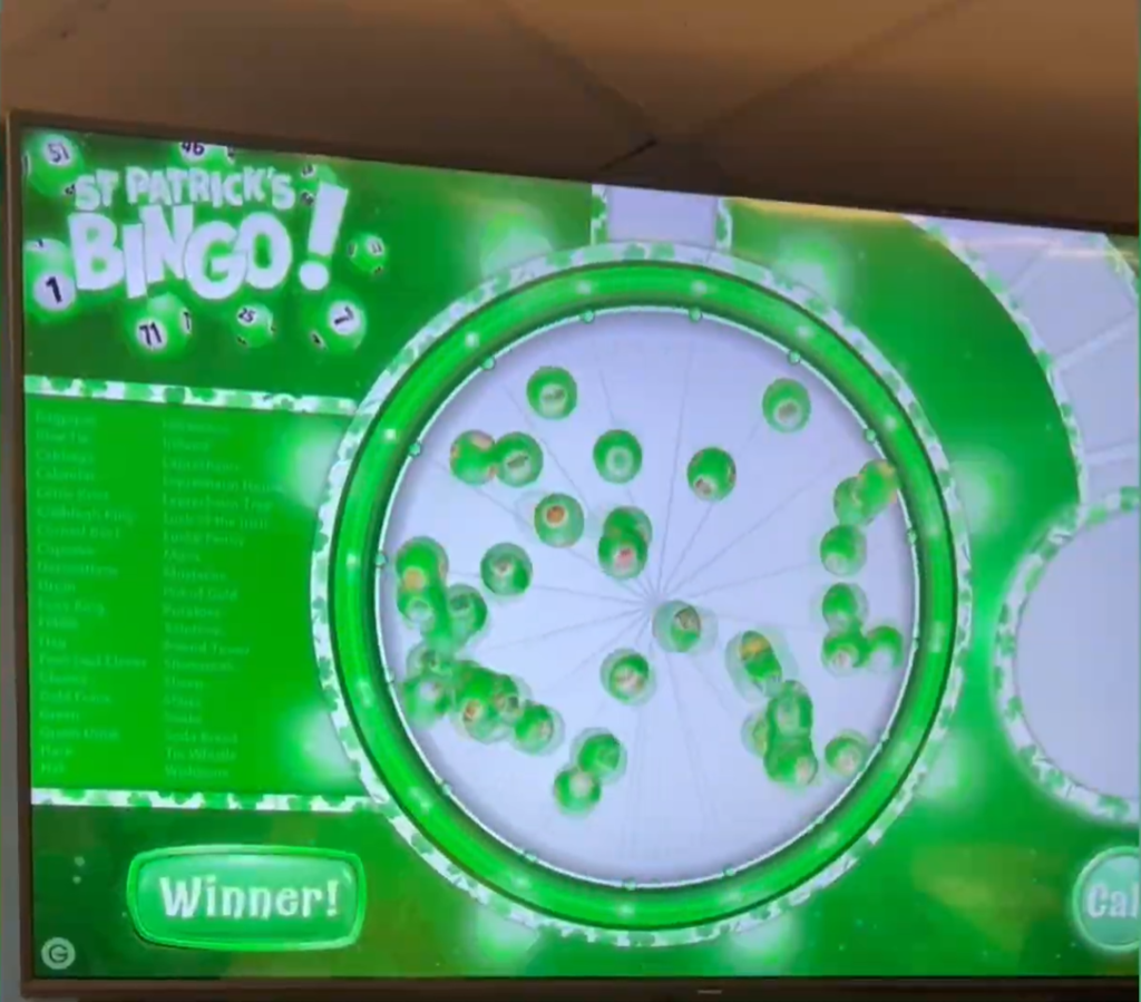 A digital display shows a green St. Patrick’s Day–themed bingo game titled “St. Patrick’s Bingo!” with numbered balls, a list of callouts, and a “Winner!” button on the screen.