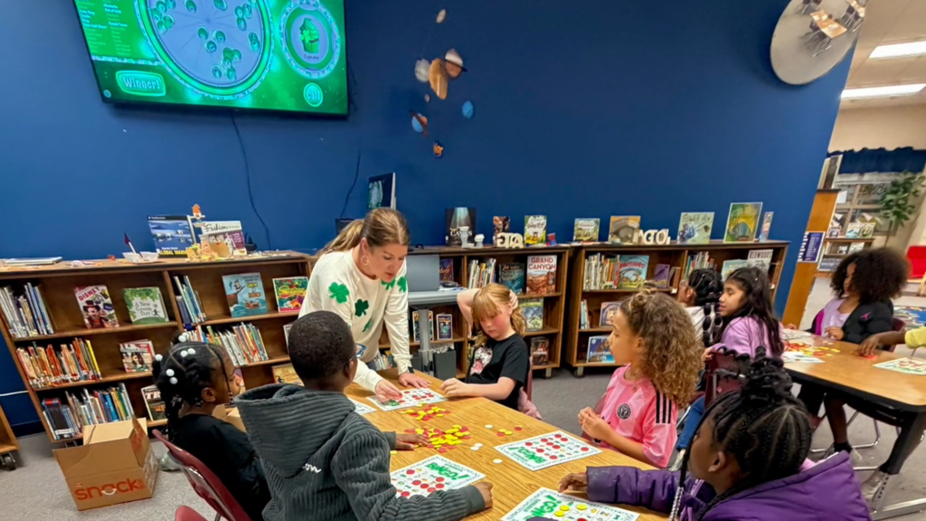 A smiling student stands in a classroom holding a popcorn bag labeled “Pop Open a Good Book.” Behind them, other children sit at a table with bingo cards and chips.