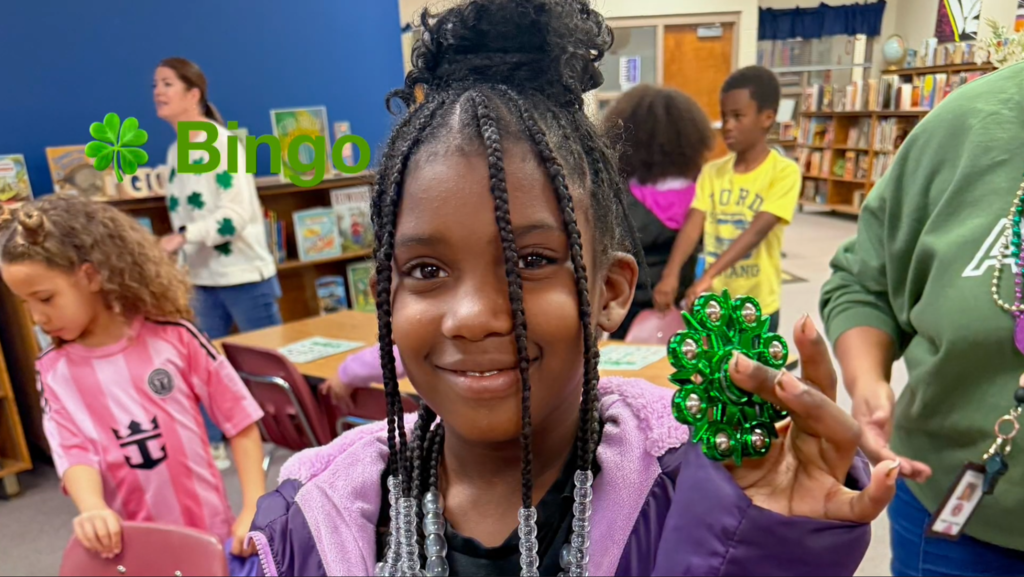 A young girl smiles while holding a shiny green shamrock‑shaped decoration with bells. She wears beaded necklaces, and other children and a teacher are visible behind her in the library. A “Bingo” graphic with a shamrock appears on the image.