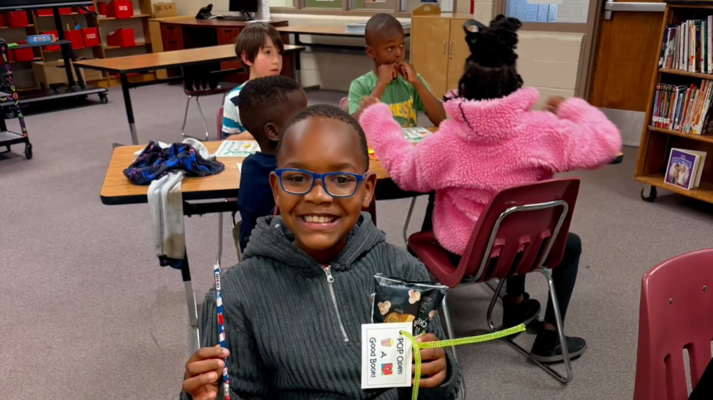 A smiling student stands in a classroom holding a popcorn bag labeled “Pop Open a Good Book.” Behind them, other children sit at a table with bingo cards and chips.