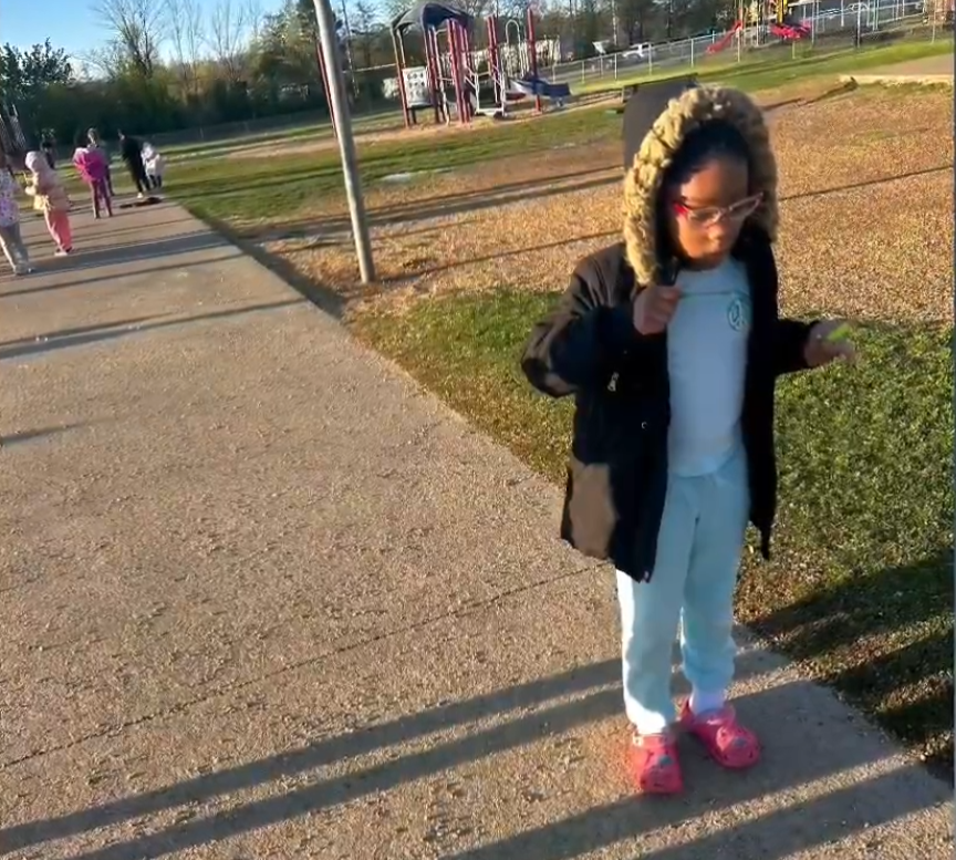A child in a black coat with a fur‑lined hood stands on a paved path holding a small object. Other children walk and play behind them near a playground.