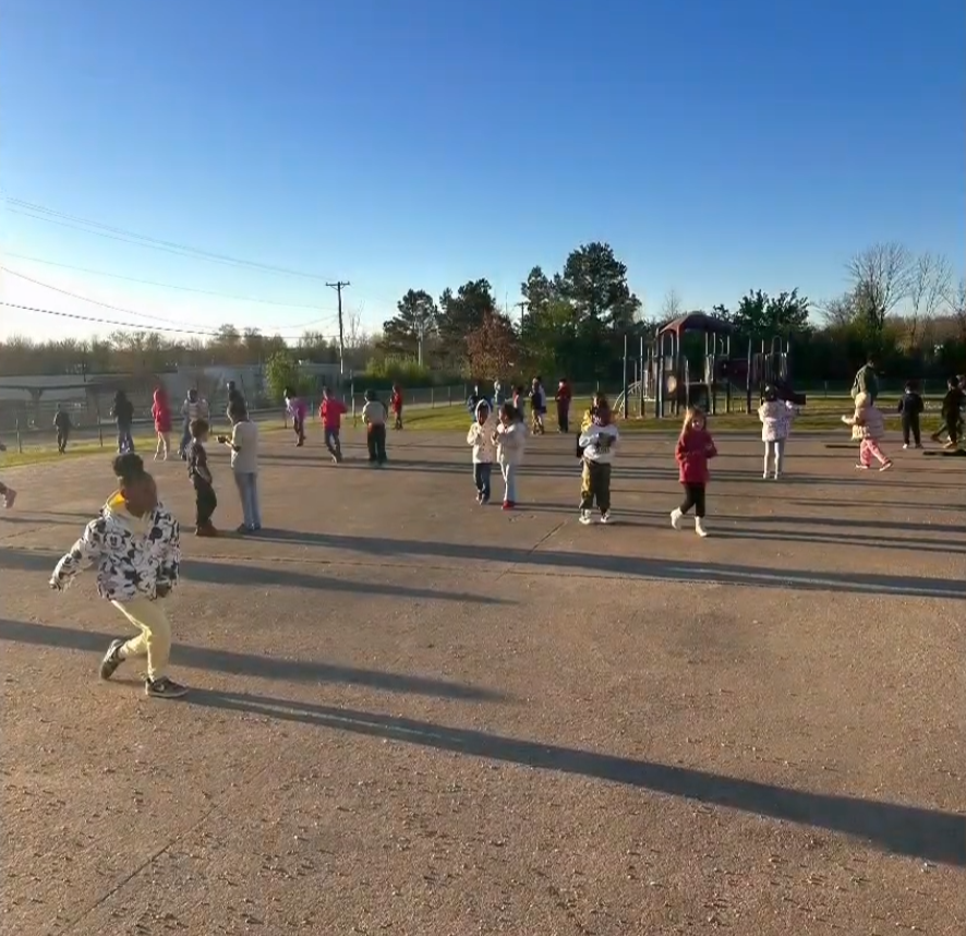 A large group of children play outside on a paved area near a playground. Students in warm clothing run, play, and enjoy the sunny weather.