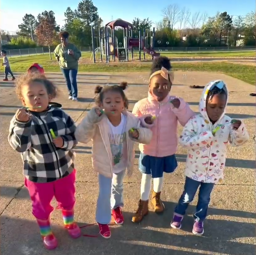 Four young children stand in a row outdoors, each blowing bubbles. A playground and supervising adult are visible behind them.