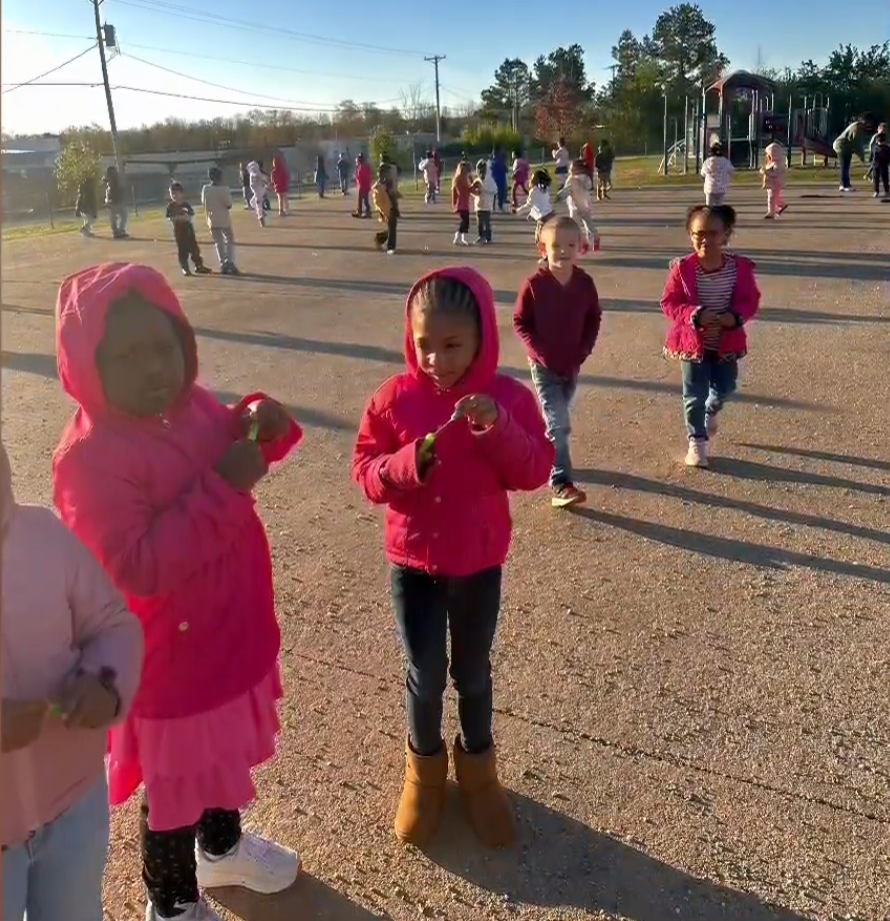 Children in bright jackets play outside on a paved area. Some hold small green items while others run and play near a playground structure.