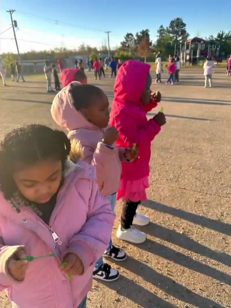 Children bundled in jackets stand outside on a sunny day blowing bubbles with bubble wands. More students and adults play in the background near a playground.