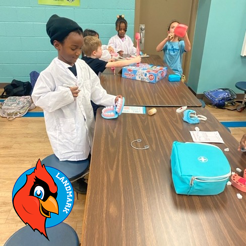Children gather around a table for a dental health activity. One child in a lab coat uses a toothbrush on a model mouth. Dental kits and tools are spread across the table. A red cardinal logo labeled “LANDMARK” is visible.