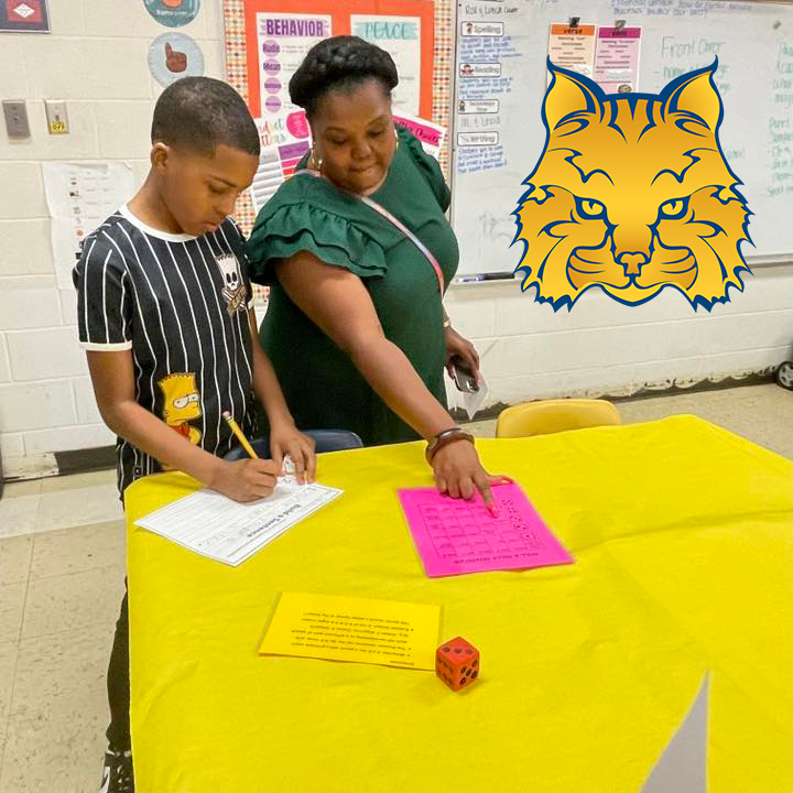 A student and adult work together at a table covered with a yellow cloth. They use dice, worksheets, and a pink grid sheet for a multiplication activity. A wildcat mascot graphic is superimposed on the image.