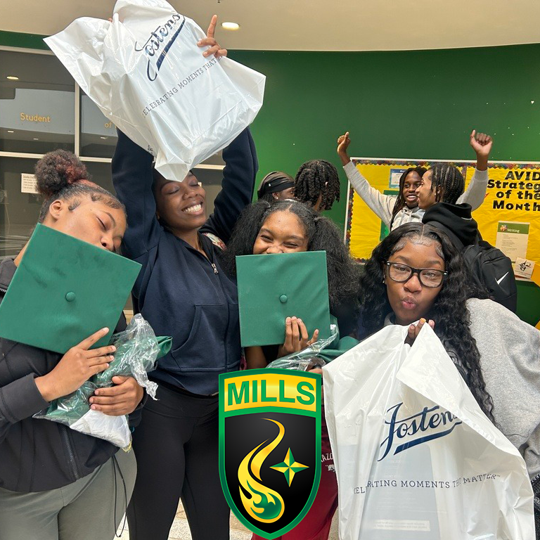 Four students smile and hold green graduation caps and Jostens bags in a classroom. A bulletin board behind them reads “AVID Strategies of the Month.” The Mills logo with a flame and star is visible at the bottom.