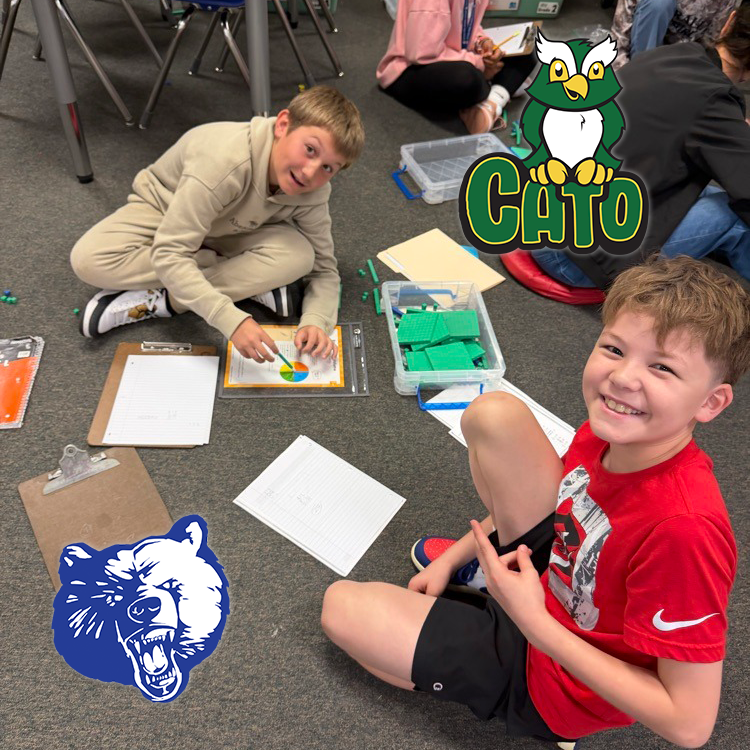Two children sit on the classroom floor working with base ten blocks, a pie chart, and clipboards. One child points at the chart while the other smiles. Logos of a bear and an owl labeled “CATO” are visible.