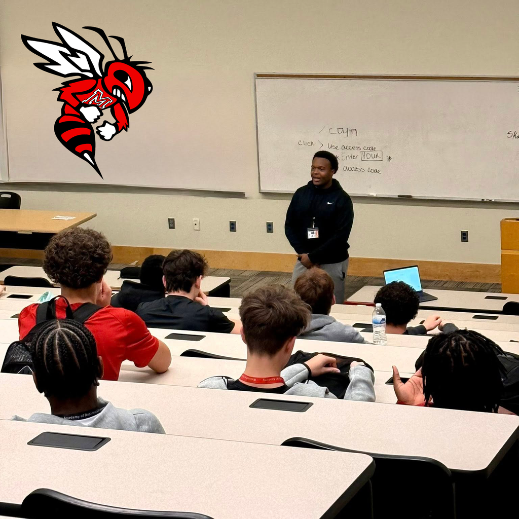 A speaker stands in front of a group of students seated in tiered rows. A whiteboard and laptop are visible behind the speaker. A large hornet mascot graphic wearing a jersey with “M” is displayed on the wall.
