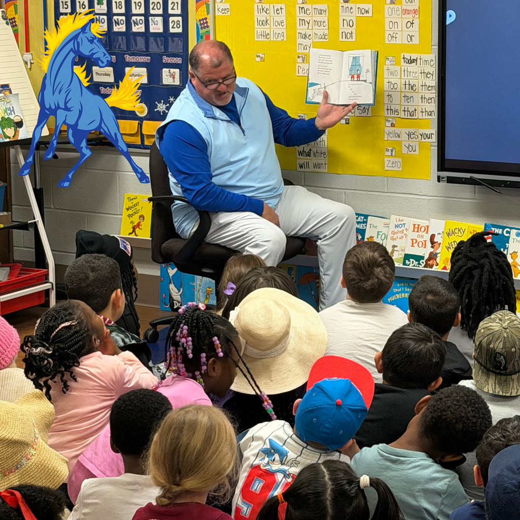 A man in a blue vest reads a book aloud to a group of young children seated on the classroom floor. The walls are decorated with sight words, a calendar, and Dr. Seuss books. A blue horse mascot graphic is visible on the left.
