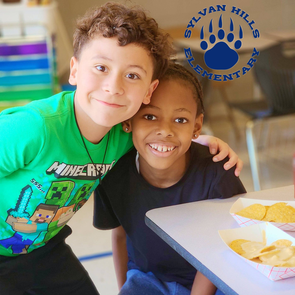 Two children sit side by side at a table, smiling at the camera with trays of nachos in front of them. The Sylvan Hills Elementary logo with a bear paw is visible in the corner.