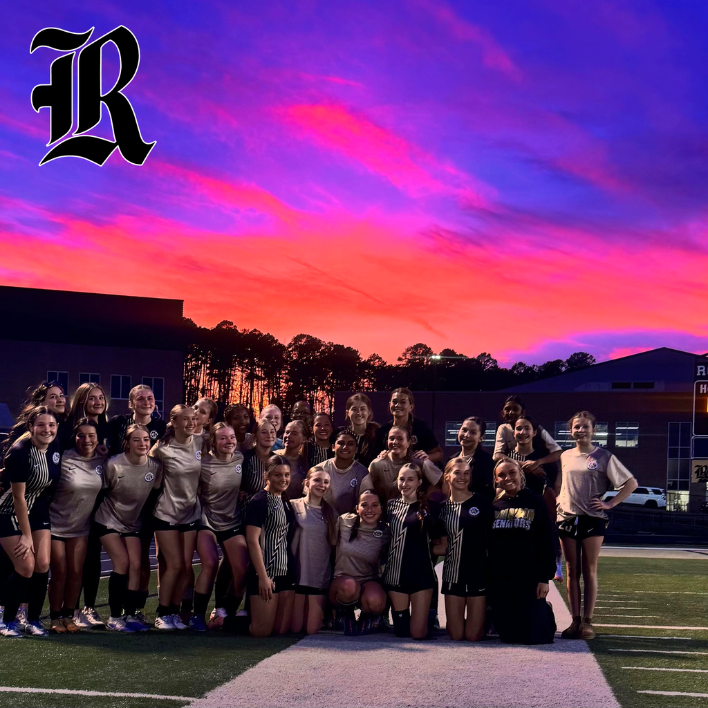 A girls soccer team poses together on a sports field during sunset. The sky glows with pink and orange hues. The background includes a school building labeled “RHS Home of the Senators.”