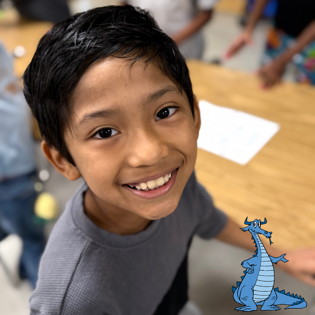 A young student smiles at the camera in a classroom, standing near a wooden table with a sheet of paper. A friendly cartoon blue dragon is illustrated in the corner, pointing playfully.