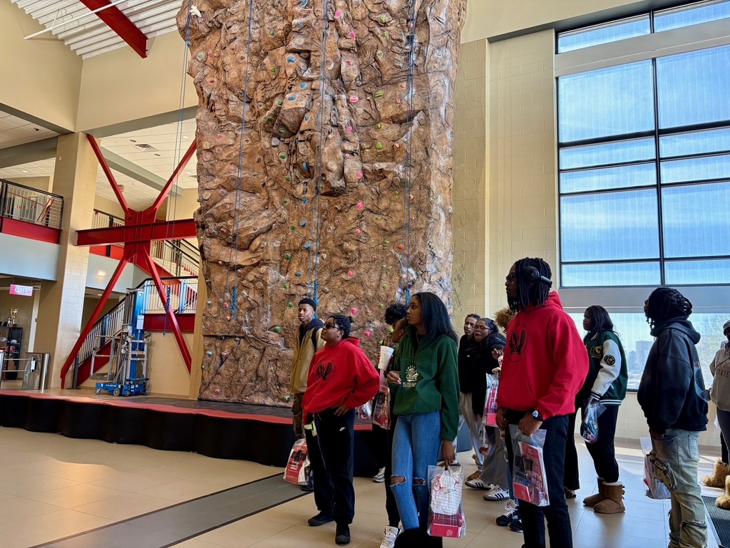 A group of students stands in front of a tall indoor rock climbing wall with ropes and colorful holds. The facility has high ceilings, red beams, and large windows.