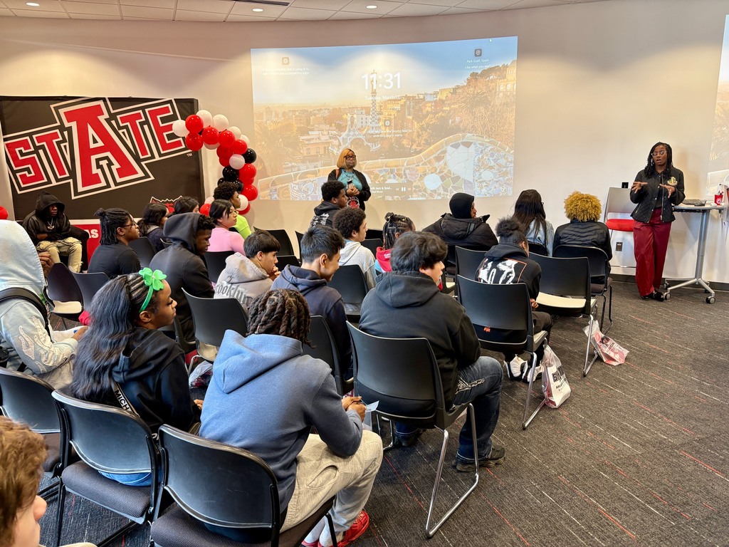 Students sit in rows facing a presenter near a screen labeled “Club Eagle Express.” A large Arkansas State University banner and a balloon arch decorate the room. Gift bags labeled “BITE” sit under chairs.
