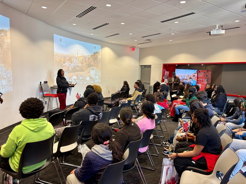 Students sit in a modern classroom listening to a speaker at the front. A large screen displays a scenic background with the time and the word “Barcelona.” A second screen in the back reads “We’re glad you are here” with a red wolf mascot.