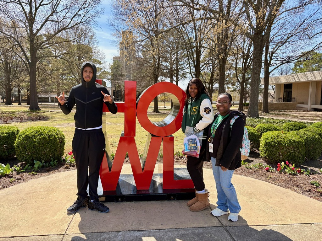 Three students pose in front of a large red “HOWL” sculpture outdoors. Trees, bushes, and a building are visible behind them on a sunny day.