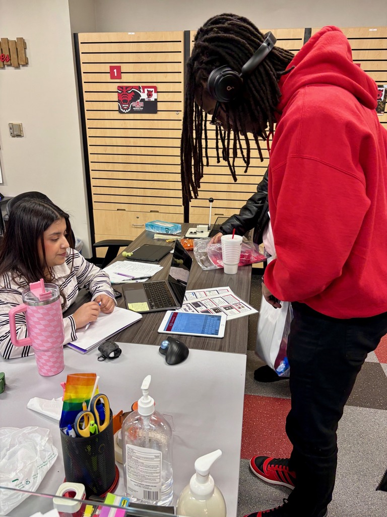 A student in a red hoodie stands at a desk speaking with another student who is seated and writing in a notebook. The desk is covered with a laptop, tablet, water bottle, sanitizer, and office supplies. A wooden wall with school logos is in the background.