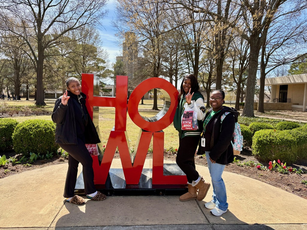Three students stand in front of the red “HOWL” sculpture, smiling and holding campus visit materials. One student holds a bag with the words “Find Your Place.”