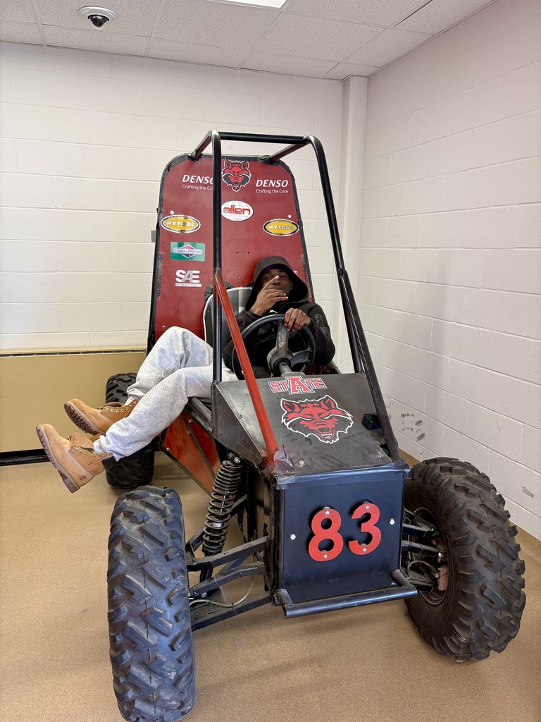 A student sits in a rugged Baja‑style engineering vehicle with large tires and a roll cage. The front displays the number 83 and the A‑State wolf logo. Sponsor logos appear on the side panels.
