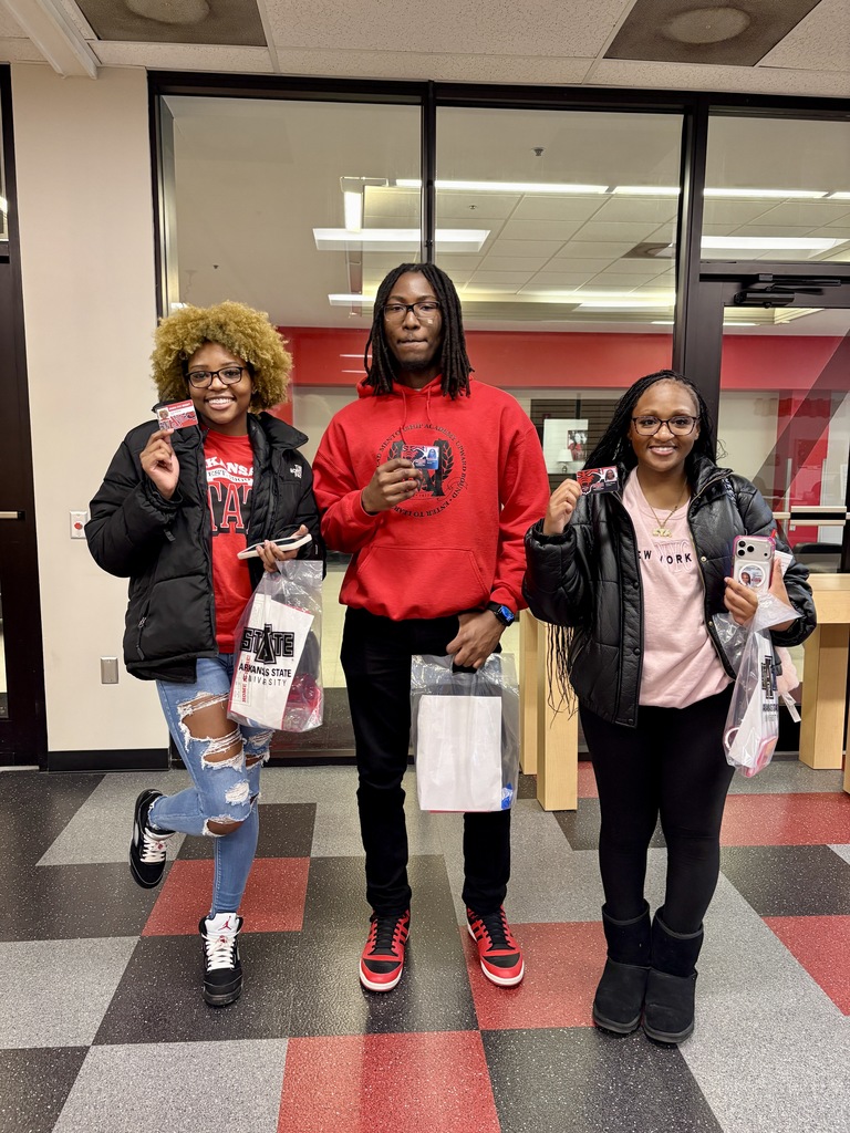 Three students stand indoors holding Arkansas State University promotional items and bags. They are smiling and standing in front of glass doors and windows.
