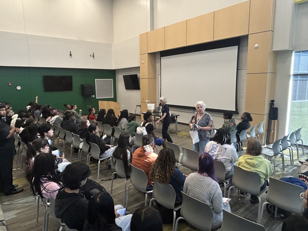 A large group of students sits in rows facing two presenters at the front of a modern lecture room. A large projection screen is behind the speakers, and bright overhead lights illuminate the space.