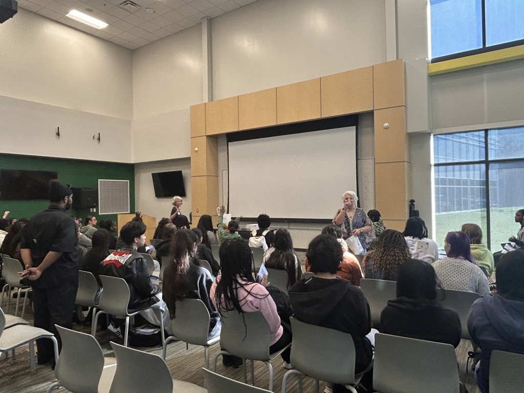 Students sit facing a presenter standing near a projection screen. One student raises a hand to participate. The room has large windows and rows of gray chairs.