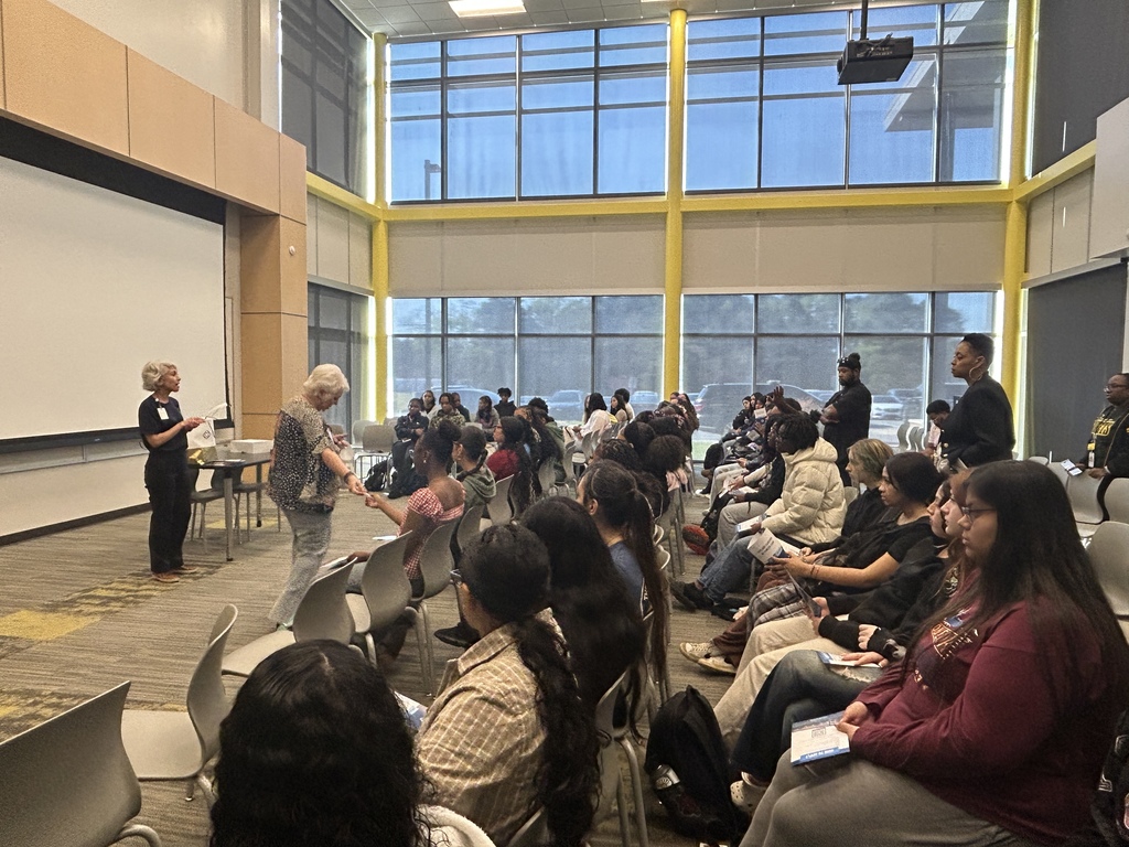 A group of people sits in rows listening to a speaker at the front of a well‑lit room with large windows. Some attendees take notes or hold materials as they follow along.
