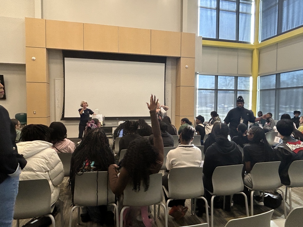 Students sit facing a projection screen while two presenters speak at the front of the room. One student has a hand raised, and sunlight streams in through tall windows along the side wall.