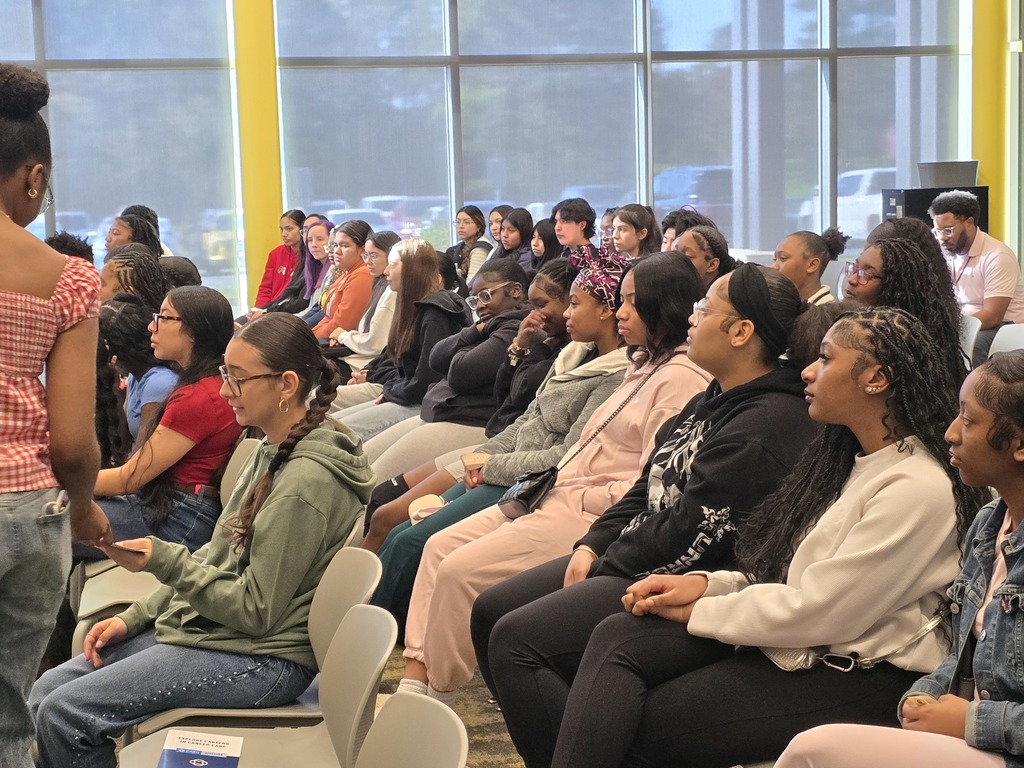 A diverse audience sits in rows inside a bright room with tall windows and yellow support beams. Two presenters stand at the front, one speaking while holding papers. Many attendees hold pamphlets.