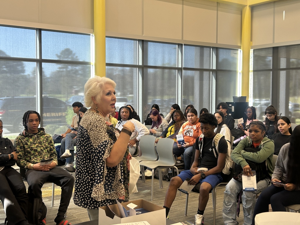 A woman stands at the front of a classroom leading a discussion with students seated in rows. Students hold informational booklets, and a sheriff’s vehicle is visible through the windows behind them.