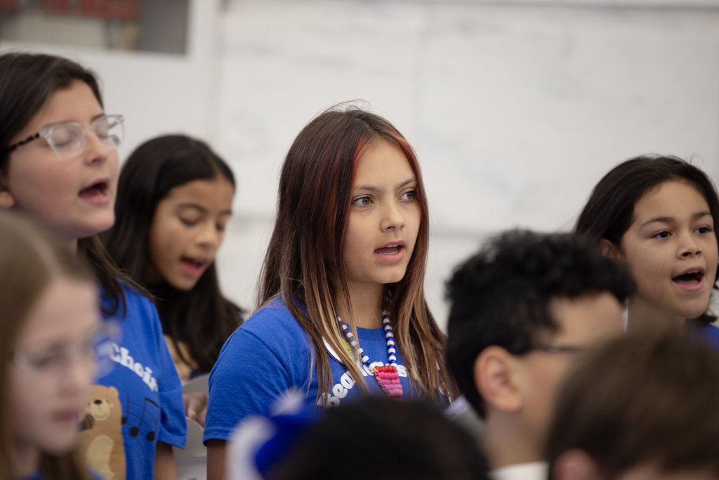 Children in matching blue “Bear Tunes Choir Sylvan Hills Elementary” shirts sing while holding lyric sheets. A teacher stands in front conducting, with a music stand between them. Adults observe from the background in a marble-walled building.